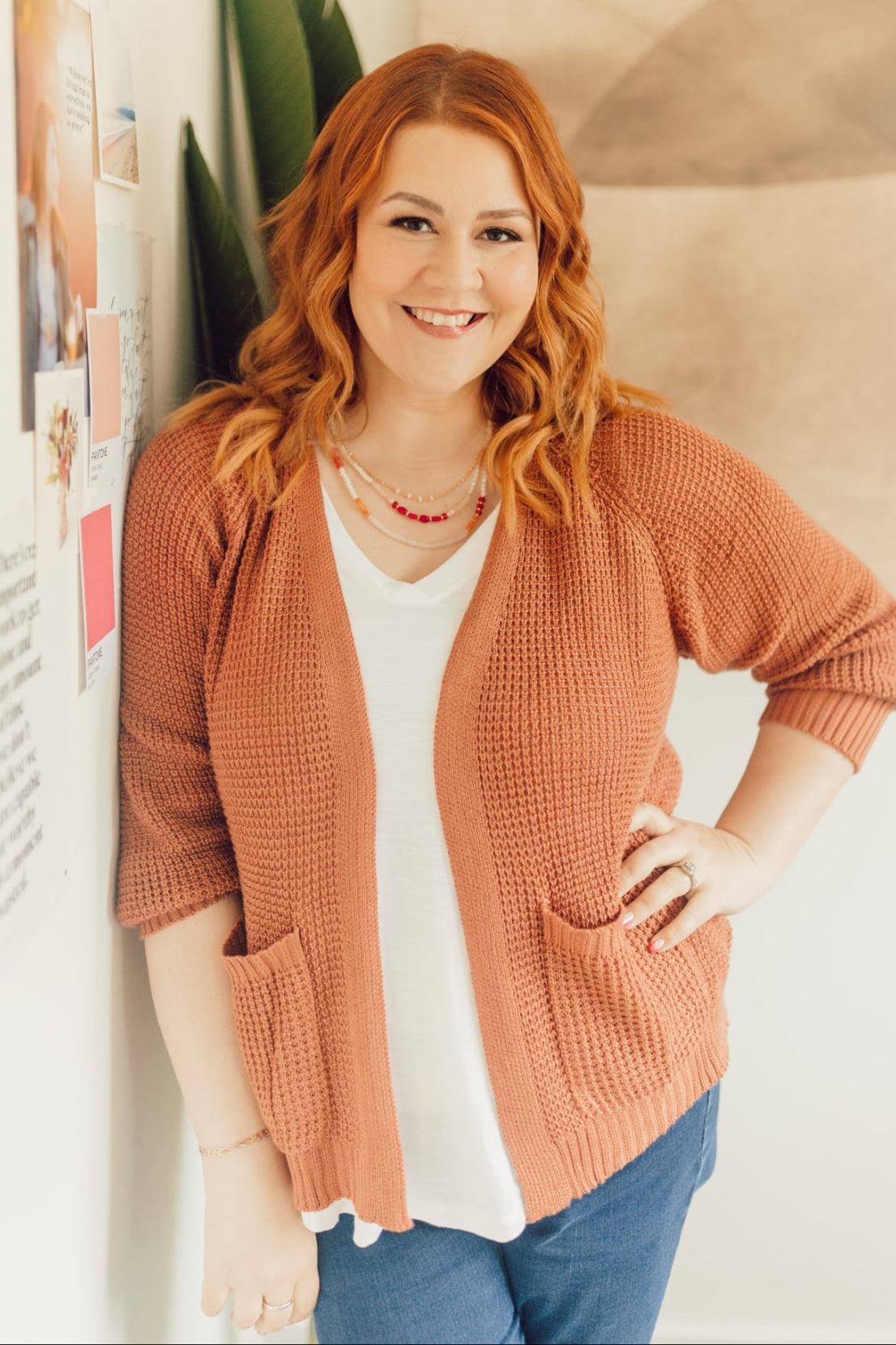Woman wearing a brown cardigan standing indoors with a plant and wall decor in the background